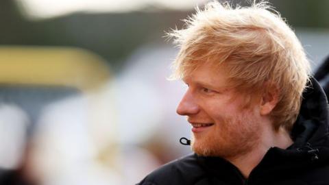 Singer Ed Sheeran smiles to the side, with an out of focus background behind him.