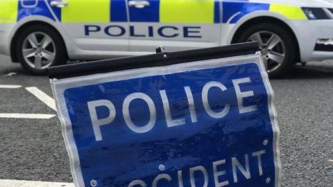 A blue police accident sign resting on the road in the foreground. In the background there is a parked police car with blue and yellow hi-vis decals.