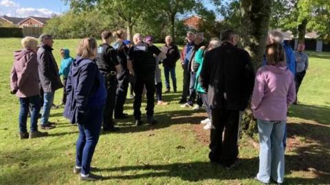 A group of people and three police officers are stood around a handful of trees on an open green space.