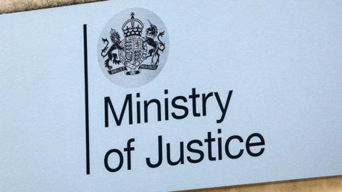 A grey Ministry of Justice sign with the words typed in black letters. The Government. The coat of arms of the United Kingdom is above, featuring a lion, unicorn and crown above some Latin words.