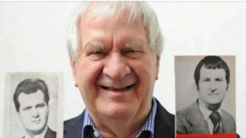 An image of Ivan Taylor who is wearing a navy suit with a blue and white checked shirt. He is holding old black and white pictures of himself as a younger man and is smiling. 