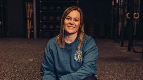 A woman with long fair hair and a blue jumper with a left chest logo is kneeling on the floor of a gym.