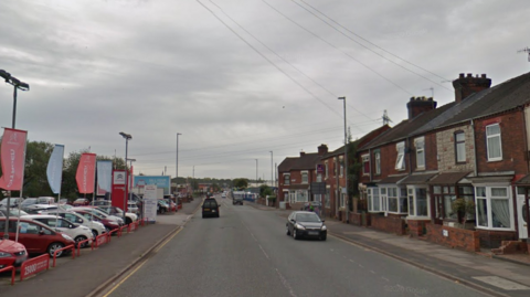 A long straight road with residential terraced housing on the right hand side. There is a car dealership on the left hand side with red and white banners on poles. There are cars driving in both directions on the road