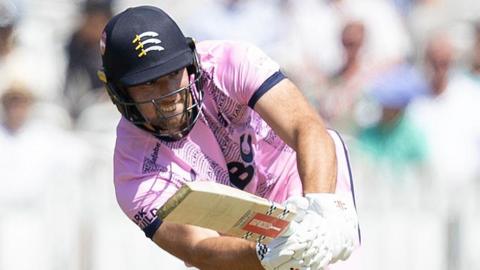 Josh de Caires batting for Middlesex holding his bat out in front of him wearing a pink shirt and helmet