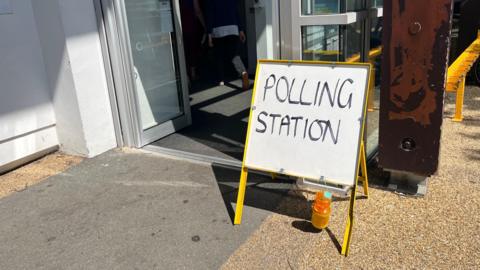 A white polling station sign is displayed on the ground next to a building. It is a white board with yellow legs. A person is walking through glass doors behind the polling sign.