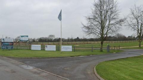 The entrance to the club, with a wooden rail fence carrying some sponsor boards, and a white flagpole with a blue flag.