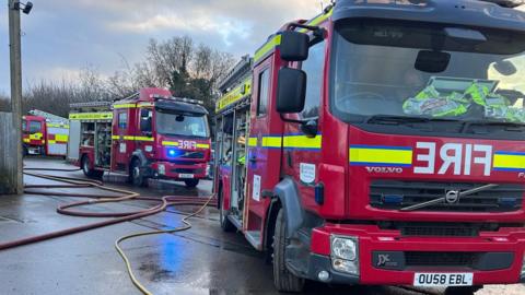 Three fire engines in a car park. Long hose reels lead off camera.