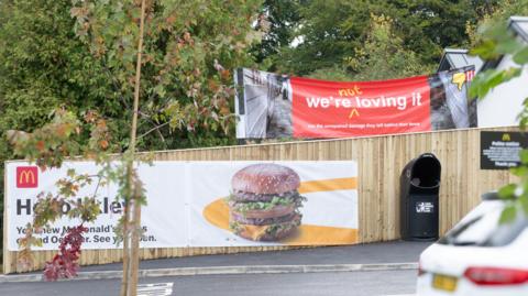 A red banner which reads 'we're not loving it' pokes up behind a wooden fence with a McDonald's advert on it. 