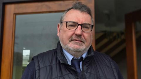 A man with grey hair, a beard and glasses with a dark blue gilet, pale blue shirt and dark blue tie standing in front of a wood framed glass door.