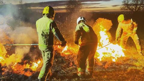 Three firefighters in full protection clothing and helmets are fighting a fire on a heath. It is just getting dark but there is still some flashes of sunlight in the sky 