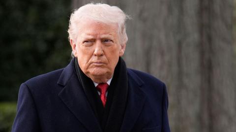 Donald Trump looks sternly to his right. He has white messy hair and is wearing a navy coat, black scarf, white shirt and red tie.