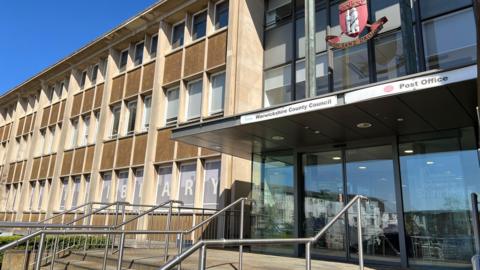 Exterior shot of a post-war council headquarters with rows of windows separated by brown render, and a more modern metal-and-glass main entrance.