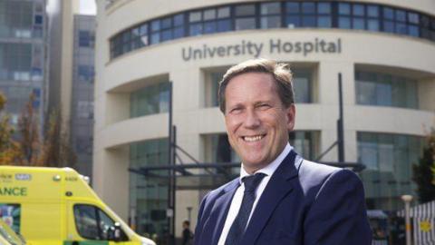 Professor Andy Hardy in front of ambulances and a hospital building made out of cream stone and glass