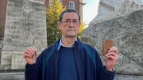 David Balfour with his father Robert's Army-issue silver-coloured fork and brown-coloured Bible. He has short, dark hair and wears glasses and a blue jacket and jumper. He is standing in front of the Hull war memorial   