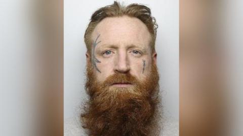 Police photo of Gavin Smedley who has a long, wiry ginger beard and hair. He has two tattoos on each side of his face.