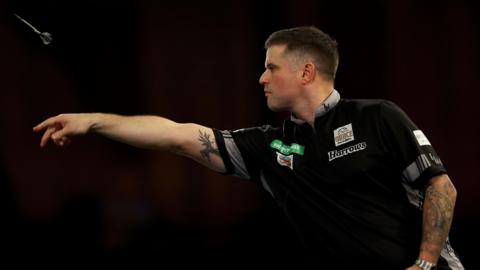 A darts player with cropped brown hair and tattooed arms wears a black top with sponsor logos on it. The background is black. His arm is outstretched having just launched a dart which is visible in the top right of the shot.