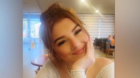 Demi-Leigh Davies smiling with her head to the side and chin resting on her hand. She is wearing a cream top and pearl bracelet and necklace and brown hair in a cafe.