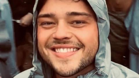 A young man with dark hair and a short beard stands smiling at the camera. he is wearing a grey raincoat with the hood pulled up. 
