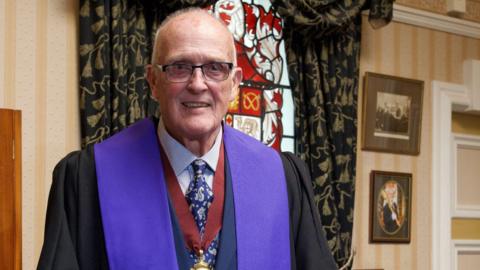 An elderly bald man with glasses, wearing a large purple sash over both shoulders, over a suit and with a ceremonial medallion around his neck, stands and smiles.