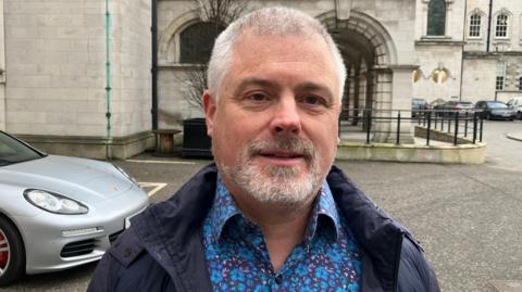 Michael Long smiles at the camera. He is wearing a shirt with blue and purple flowers and a navy coat. He has grey hair and a grey beard.