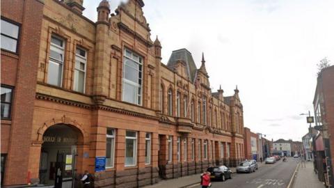 A red brick two-storey building with gables along the front. Cars are parked on the road outside.
