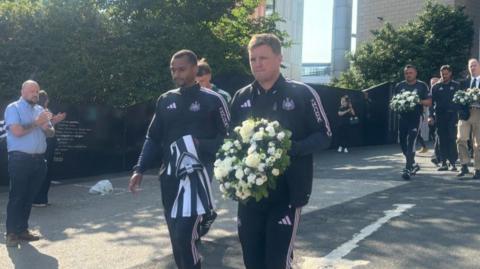 Newcastle United head coach Eddie Howe walks with club players and staff carrying a floral wreath of white and cream flowers with green foliage for Kevin Keegan at St James' Park. Several bystanders clap as he walks by.