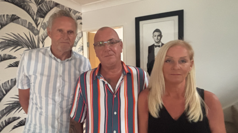 A grey-haired man in a grey and white vertical striped shirt, a bald man with a grey, orange and white vertical striped shirt, and a woman with long blonde hair and a black sleeveless top