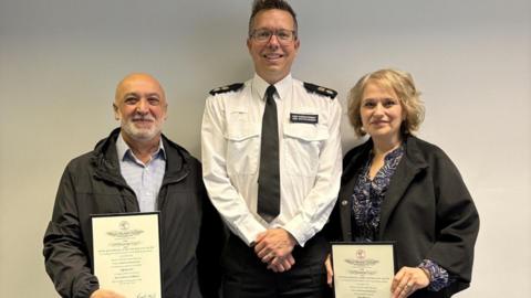 Luigi wearing a black jacket and striped shirt holding his certificate to standing next to Jon Hutchinson in his police uniform of white shirt and black trousers. Tina is standing on the police officer's left with her certificate, wearing a black coat and a blue printed shirt- all three smiling for camera.