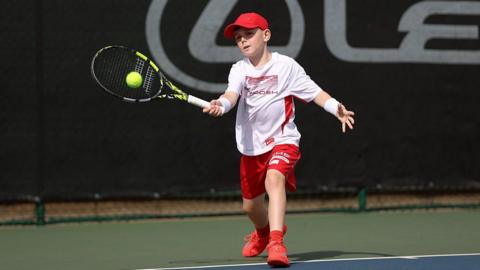 A boy is playing tennis on an outdoor court, holding a racket and striking a bright yellow ball. The person is wearing a red cap, a white sports shirt, red shorts, and red athletic shoes. A dark backdrop with large lettering is visible behind the court, highlighting the action in the foreground.