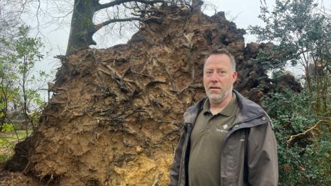 Gary Long stands in front of a huge root ball of a tree which has fallen. He looks serious. He has short brown and grey hair and a grey beard. He is wearing a Trewithen-branded top under a wax jacket.