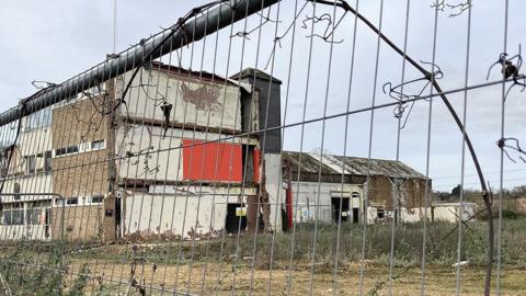 A view of a derelict factory through a silver metal fence. The building is partially demolished with broken windows and is surrounded by overgrown grass and weeds.