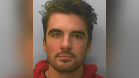 Police custody photograph of a man with brown hair and stubble wearing a red hoodie