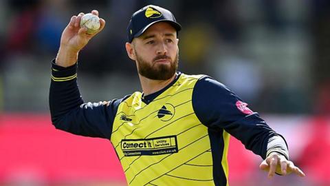 James Vince shapes to throw a white cricket ball, dressed in Hampshire Hawks' yellow shirt with thin, navy blue diagonal stripes and navy blue sleeves