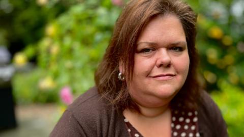 Caroline Fonjock looks at the camera. She is wearing a brown spotty dress and a chocolate brown matching cardigan. She has dark brown shoulder-length hair and silver hooped earrings on. She is standing in her garden with a yellow rose bush blurred in the background and some shrubbery to her right.