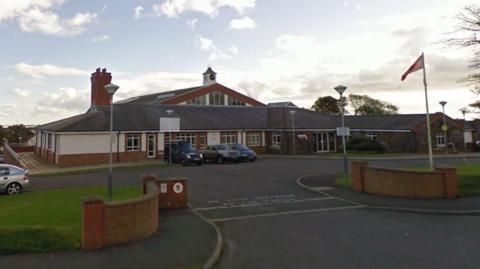 The exterior of Cronk y Berry school, a driveway leads to the large brown and white building which has a clock on the roof. Cars are parked in front of the building, a Manx flag flies on a flagpole on the grounds.