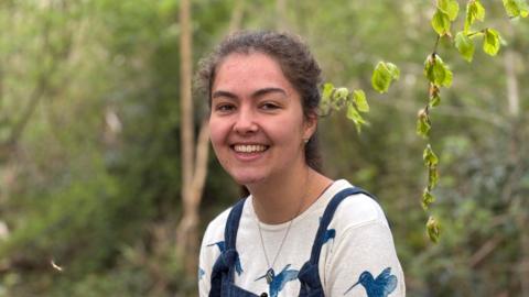 A young woman with her hair in a ponytail smiling at the camera. She's sitting on a tree stump in woodland at Moseley Bog in Birmingham and is wearing a white top with blue birds on it underneath a pair of blue corduroy dungarees. 