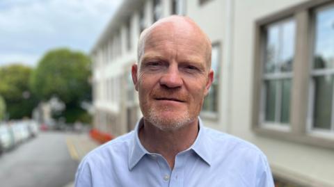 Deputy Gavin St Pier - A man with a bald head looking at the camera. He has a ginger and grey beard, and is wearing a blue button-up shirt with the collar open. 