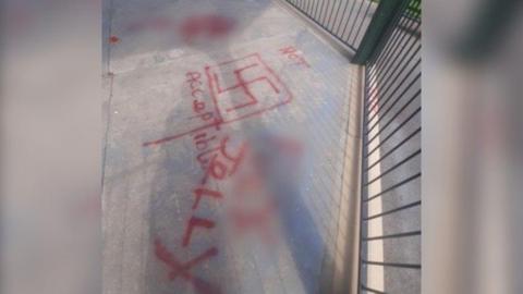Grey ground surrounded by railings of a bandstand. You can see red spray-painted images, with a swastika symbol with a red square around it, as well as the words 'not acceptable' and 'jolly'. Part of the spray paint is blurred out.