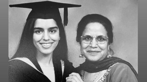 A black and white image of Manj Kaur who has long dark hair and is wearing a black mortarboard and a gown. She is standing next to her mum who has dark hair tied back and is wearing glasses and a dupatta scarf around her neck