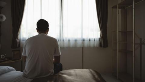 Man facing away from the camera looking towards a window. He is sitting down on a bed in a bedroom