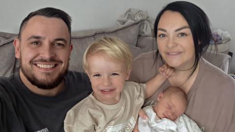Man with short dark hair and beard with a dark top with a child with blonde hair and a beige top, a woman with brown hair and a brown top and a baby wrapped in white. They are sitting on a sofa.