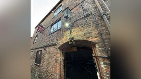 An old gatehouse in a town centre, a three-storey brick building with a large arch. 