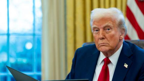 US President Donald Trump stares straight ahead during an executive order signing ceremony in the Oval Office of the White House. He wears a dark blue suit and red tie and a US flag pin on his lapel. 