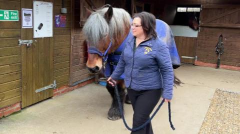 A woman leads a long-haired brown pony through a stable yard.