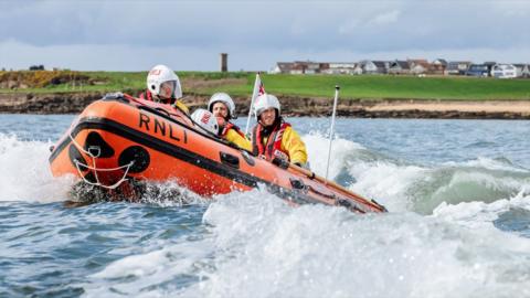 A three-person RNLI crew in yellow, wearing helmets, on board a yellow lifeboat on the sea.