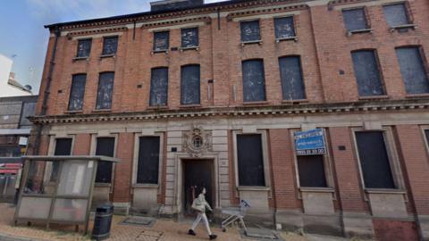 A red brick building of about three storeys, with boarded-up windows, next to a pavement. There is a discarded shopping trolley on the pavement. A person is walking past. It has the look of a Victorian factory. There is a plastic bus shelter and a bin in front of the building.