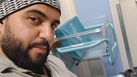 A selfie of a man who is sitting down in a hospital. He is looking directly at the camera. He is wearing a beige polo shirt and wearing a black and is wearing what appears to be a white knitted kufi hat.