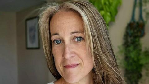 Poppy Whiteside smiling slightly at the camera with her home, with a framed piece of art and plants on the wall, in soft focus in the background