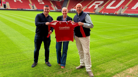 Two men stand either side of a woman on a football pitch, holding a red football shirt. 