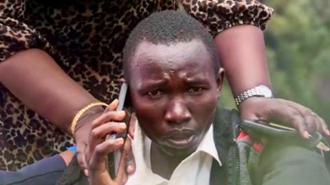 Felix Kemboi is comforted by a loved one who places her hands on his shoulders. He is visibly update as he holds a mobile phone to his ear. Image has been colour-corrected for distortion.